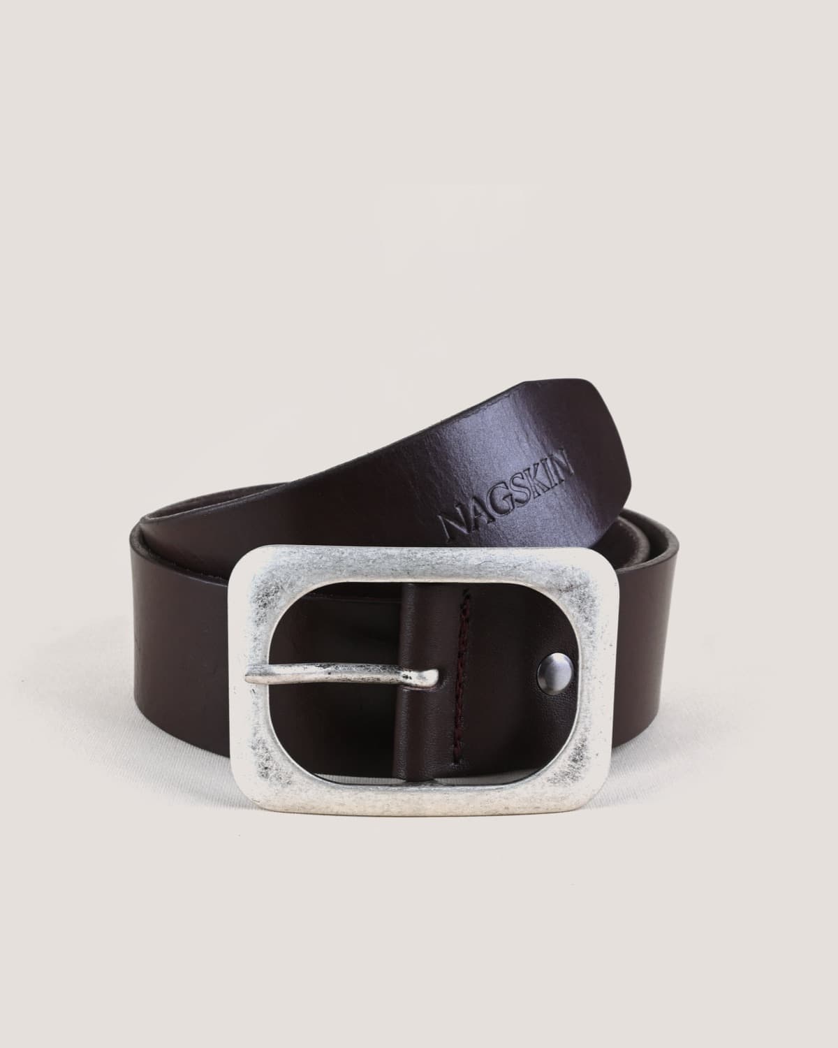 A dark brown leather belt coiled and shot head-on, NAGSKIN embossed along the strap above an antiqued silver oval buckle