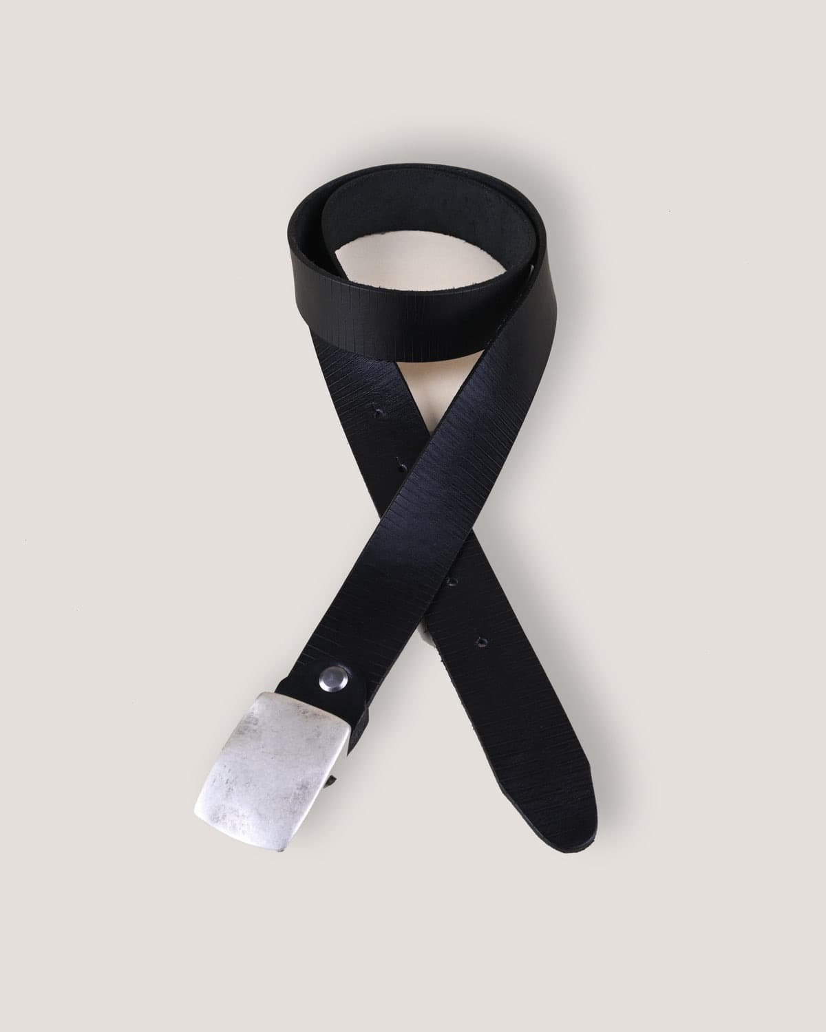 A black leather belt coiled into a loop against a bone-white backdrop, its brushed silver rectangular buckle resting at the bottom left